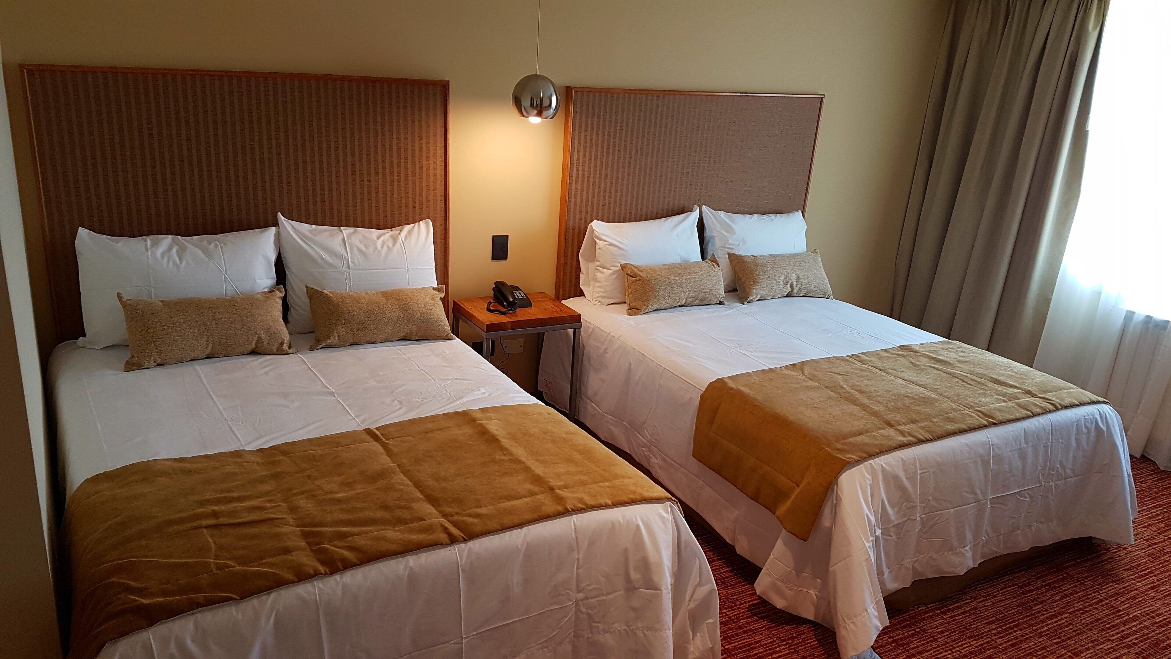 A hotel room with two beds, a bedside table with a phone, and a window with curtains.