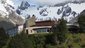 A modern building with large windows and a stone facade, surrounded by lush greenery and snow-capped mountains in the background. - Las Lengas Hotel (Ushuaia)
