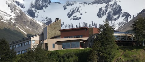 Een modern gebouw met grote ramen en een stenen gevel, omgeven door weelderige begroeiing en besneeuwde bergen op de achtergrond.