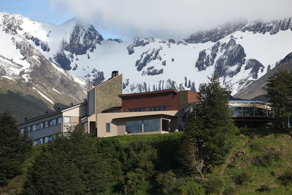 A modern building with large windows and a stone facade, surrounded by lush greenery and snow-capped mountains in the background.