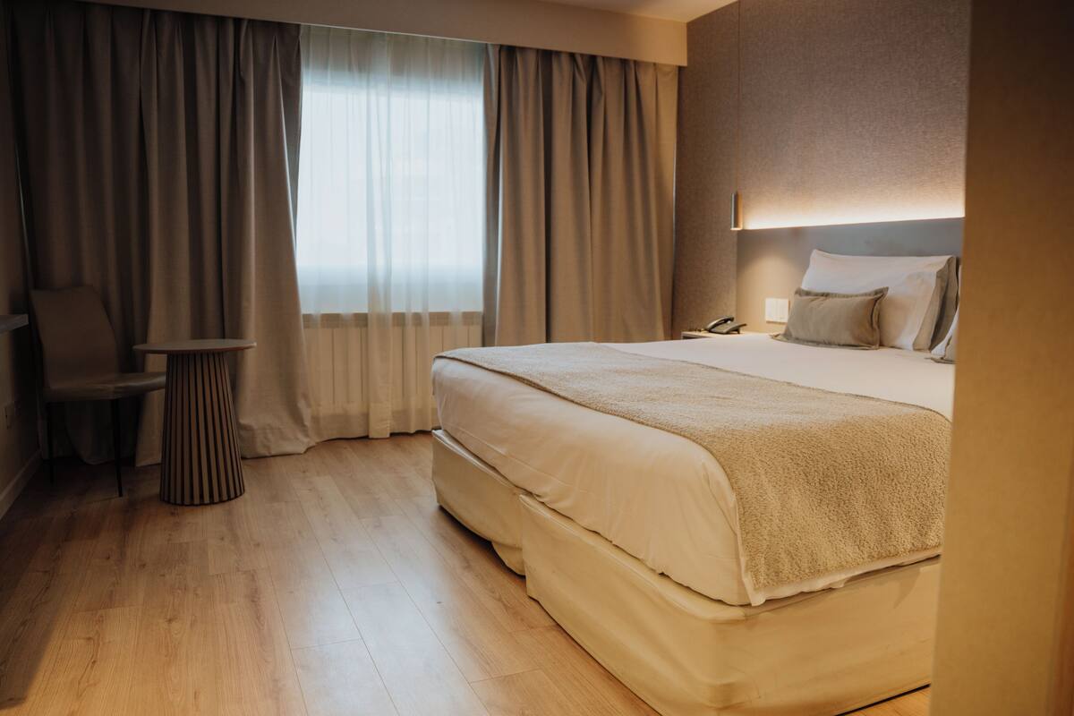 A hotel room with a large bed, a chair, a small table, and a window with curtains.