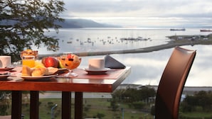 A breakfast table with croissants, fruit, and juice, overlooking a calm body of water with boats. - Las Lengas Hotel (Ushuaia)