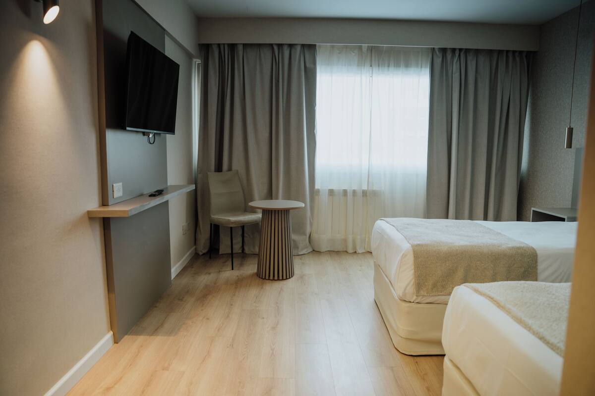 A hotel room with a bed, a chair, a small table, a TV mounted on the wall, and a window with curtains.