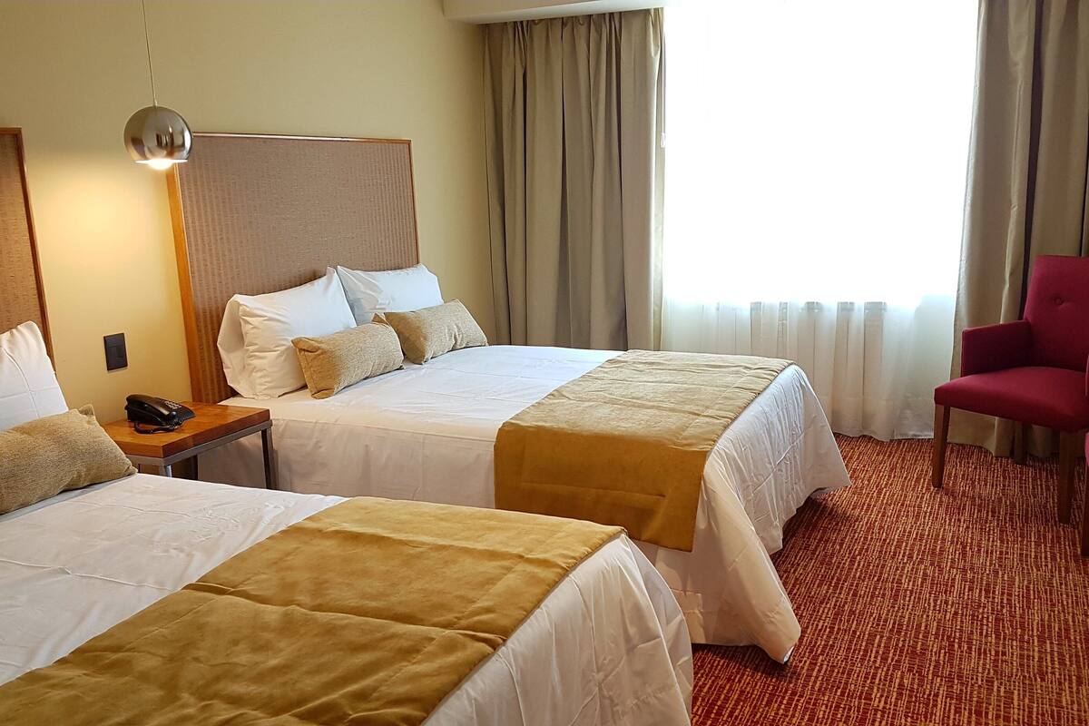 A hotel room with two beds, a desk, a chair, and a large window with curtains.