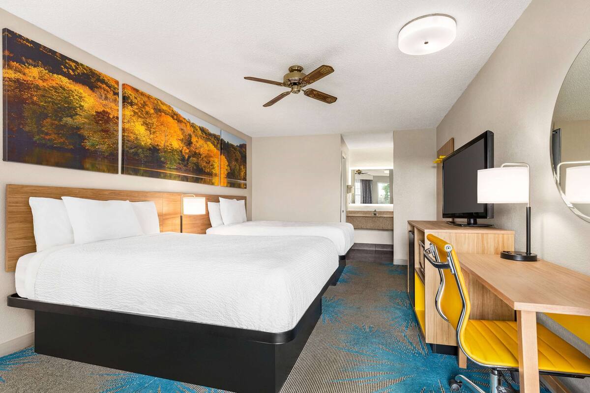 A hotel room with a bed, two bedside tables, a desk with a chair, a ceiling fan, and artwork on the wall.