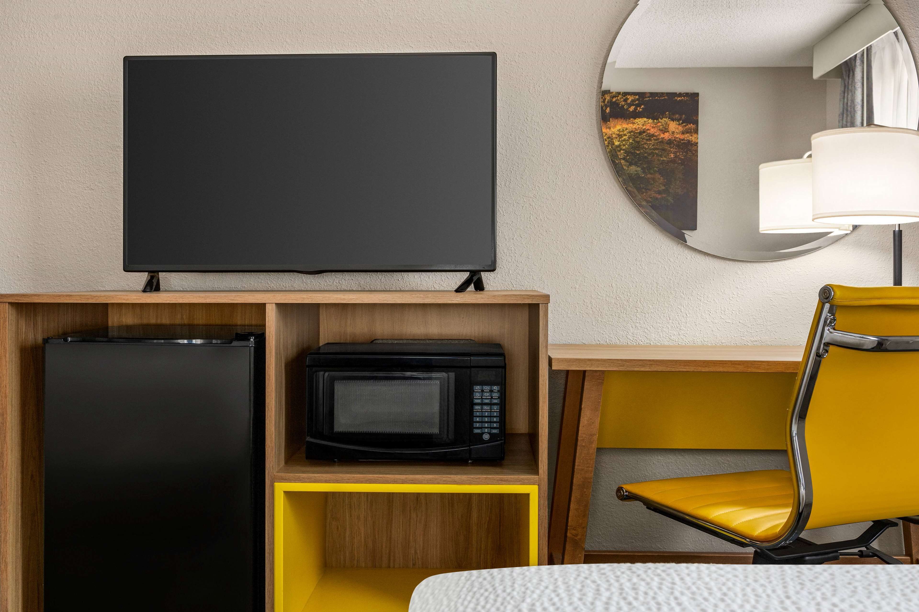A hotel room with a flat-screen TV on a wooden stand, a microwave, a refrigerator, a yellow chair, and a round mirror on the wall.
