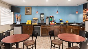 A small, well-lit kitchen area with a blue wall, wooden cabinets, a refrigerator, a microwave, a coffee maker, and a sink. There are two round tables with chairs, and a television mounted on the wall.