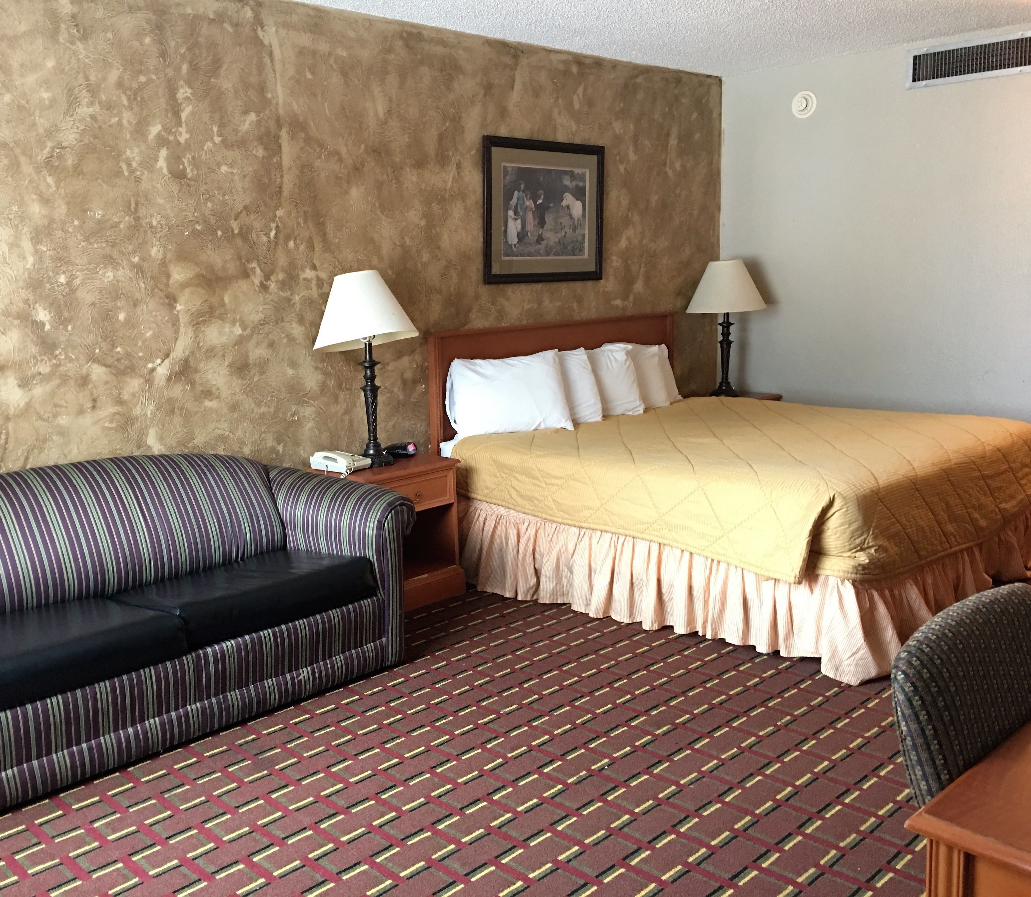 A hotel room with a bed, two bedside lamps, a telephone, a striped sofa, and a patterned carpet.