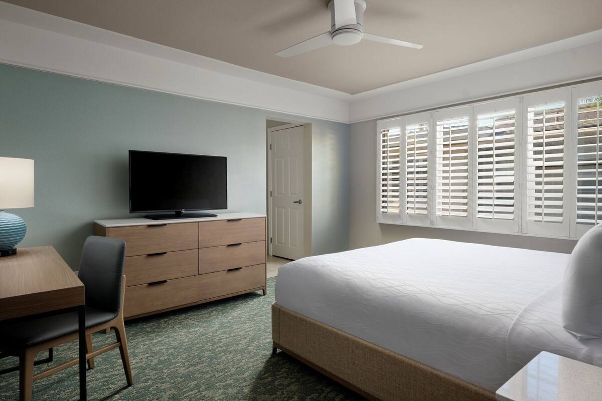 Ein Hotelzimmer mit einem Bett, einem Schreibtisch mit Fernseher, einem Sessel, einer Kommode und einem Fenster mit Jalousien.