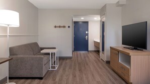 Una habitación de hotel moderna con un sofá gris, una mesita, un televisor sobre un mueble de madera y una puerta azul al fondo.