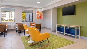Un área de espera moderna con alfombra verde, sillas amarillas, un banco de madera y un televisor en la pared.