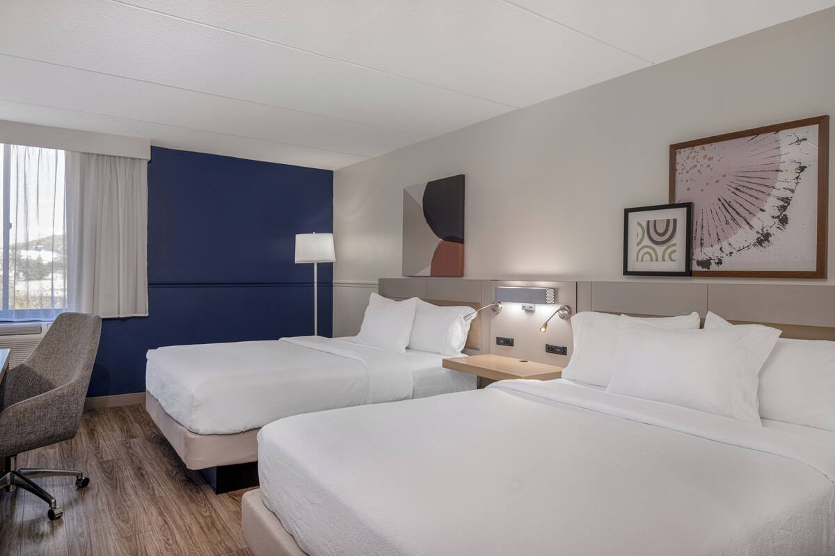 Hotellihuone, jossa on kaksi sänkyä, työpöytä, tuoli ja ikkuna, jossa on verhot.