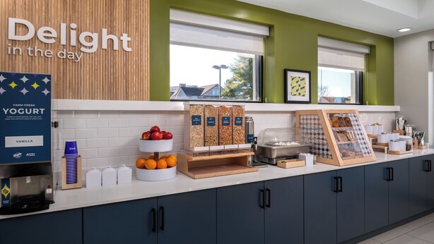 Una barra de desayuno con un letrero que dice "Delight in the day", un dispensador de yogur, cuencos de fruta y una máquina de café.