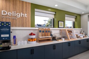 Un comptoir pour le petit-déjeuner avec une pancarte indiquant « Savourez la journée », un distributeur de yaourts, des bols de fruits et une machine à café.