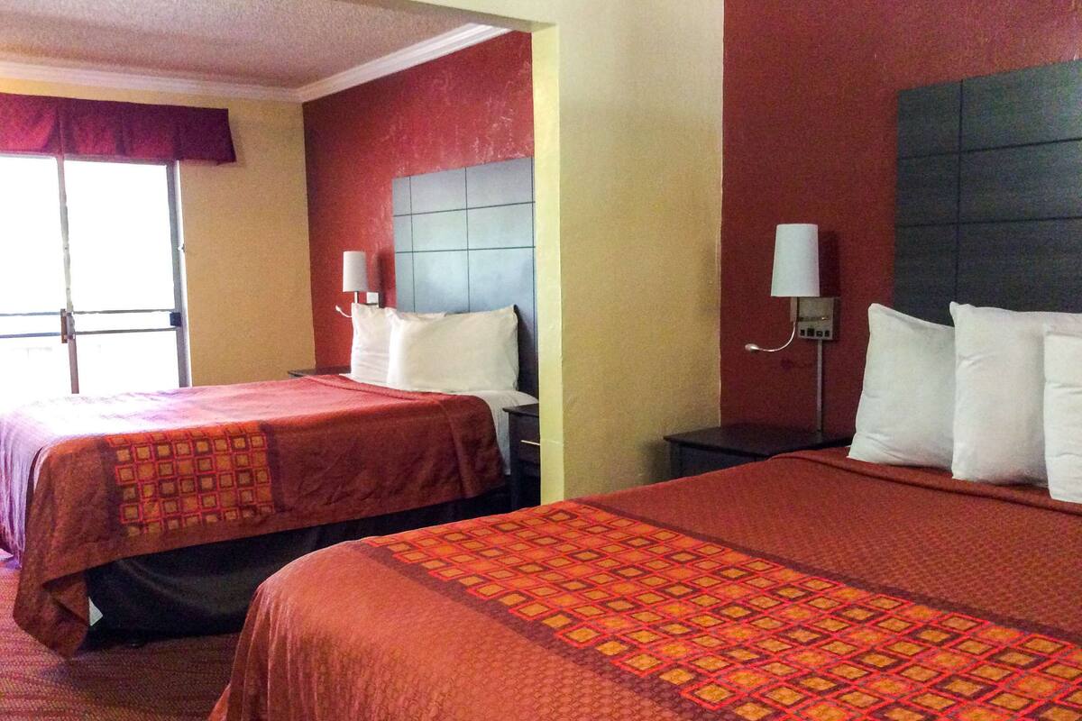 A hotel room with two beds, each with a red bedspread and white pillows. There are two bedside tables with lamps, and a window with a red curtain.