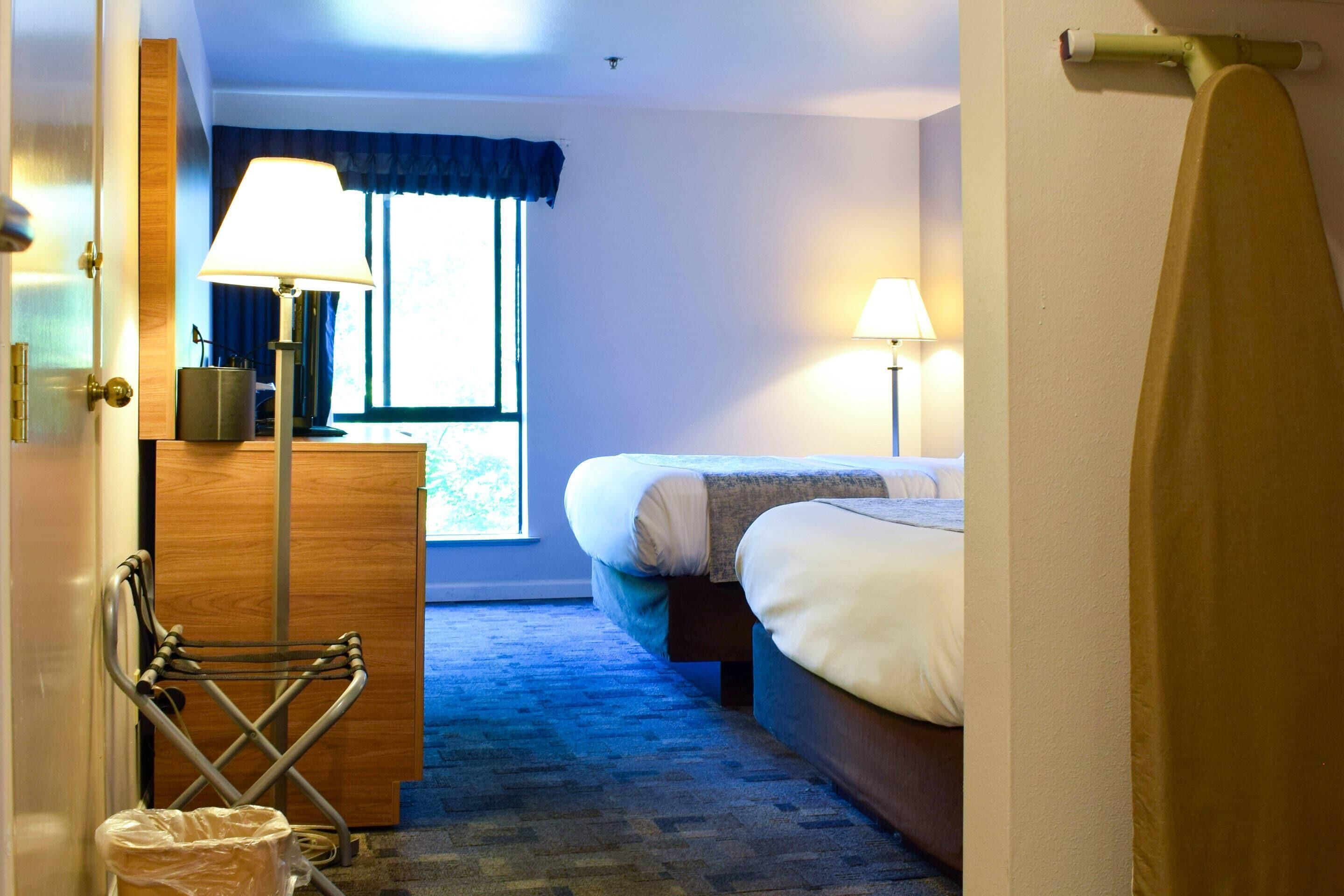 Una habitación de hotel con una cama, un escritorio con lámpara, una ventana con cortinas y un perchero para toallas.