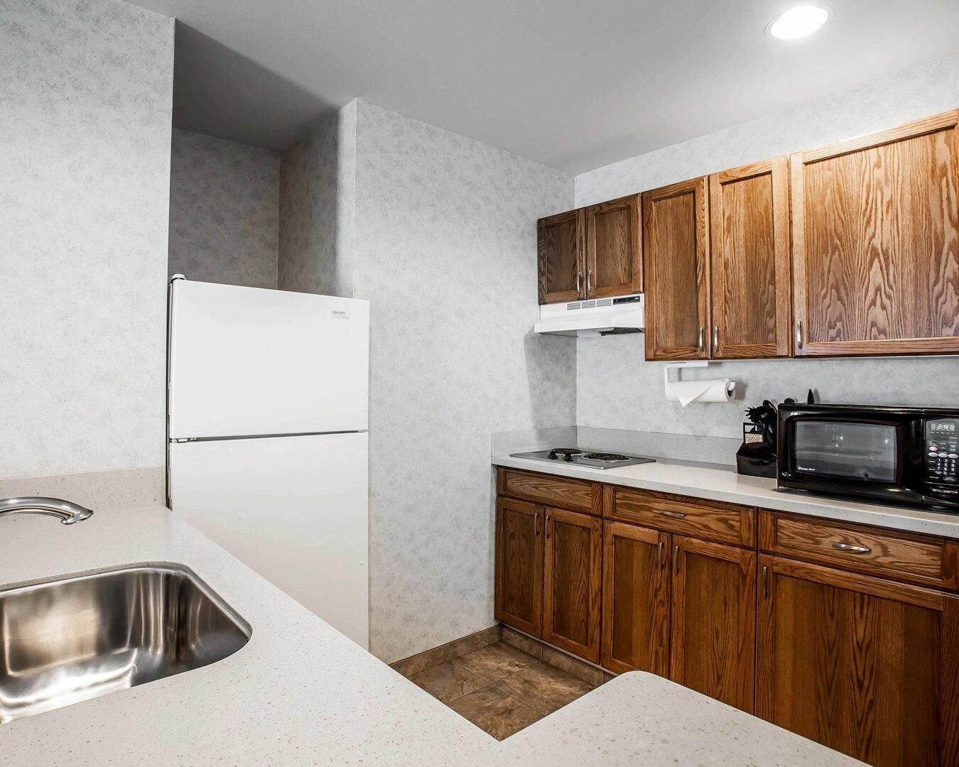 A compact kitchen with a stainless steel sink, a white refrigerator, wooden cabinets, a black microwave, and a gas stove.