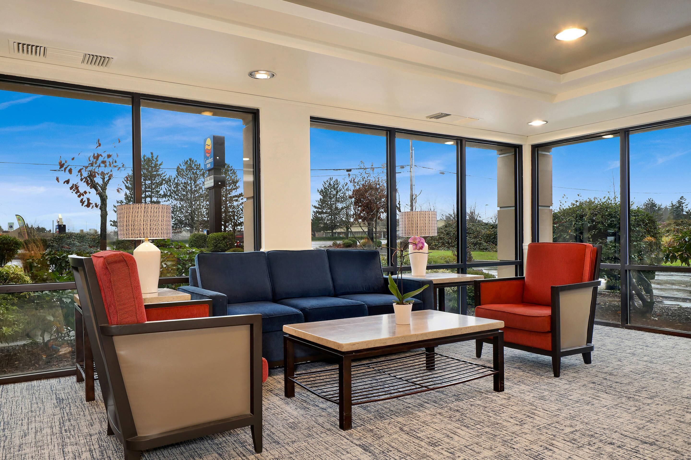 A modern hotel room with large windows, a sofa, armchairs, a small table, and a lamp.