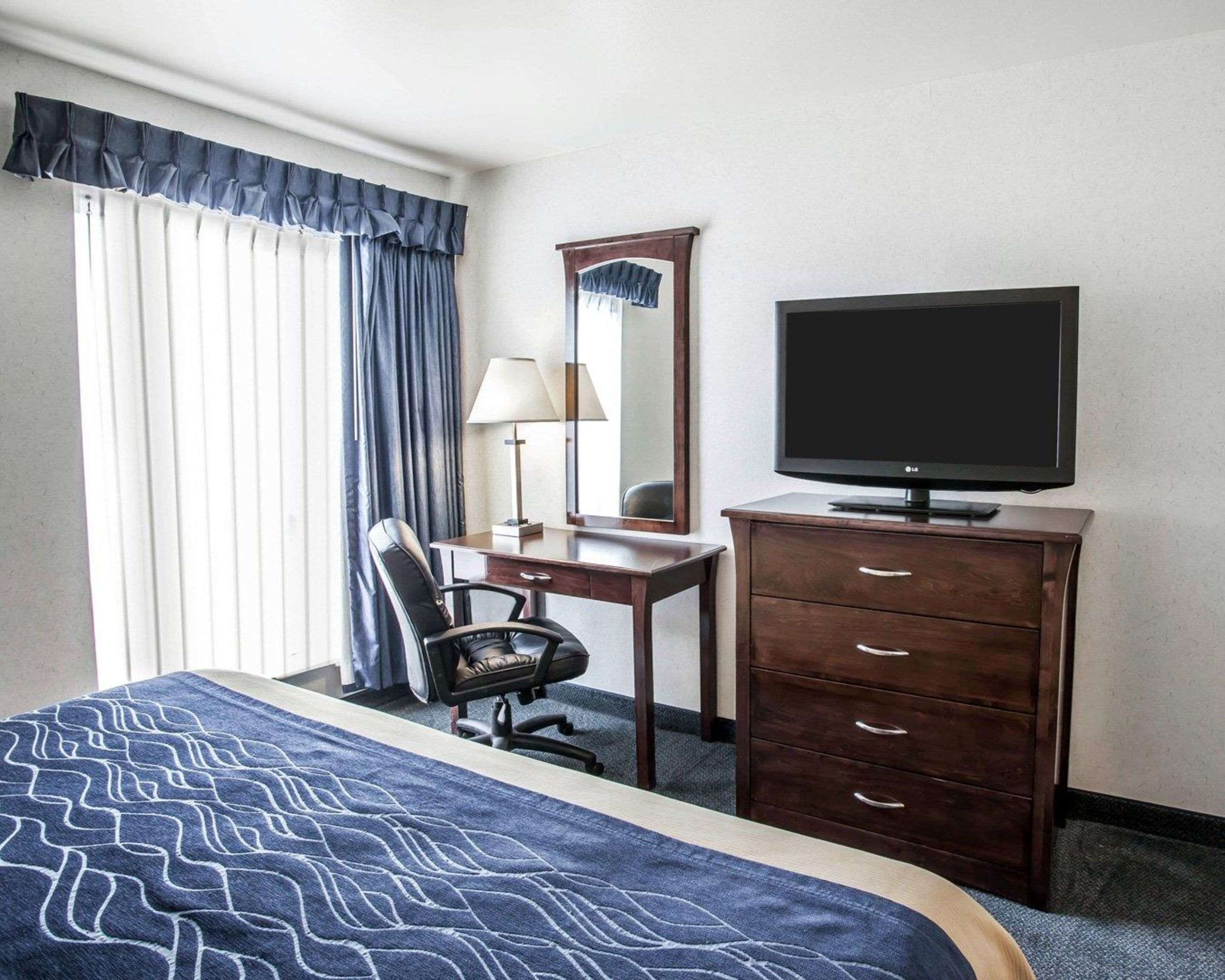 A hotel room with a bed, a desk, a chair, a dresser, a television, and a window with curtains.