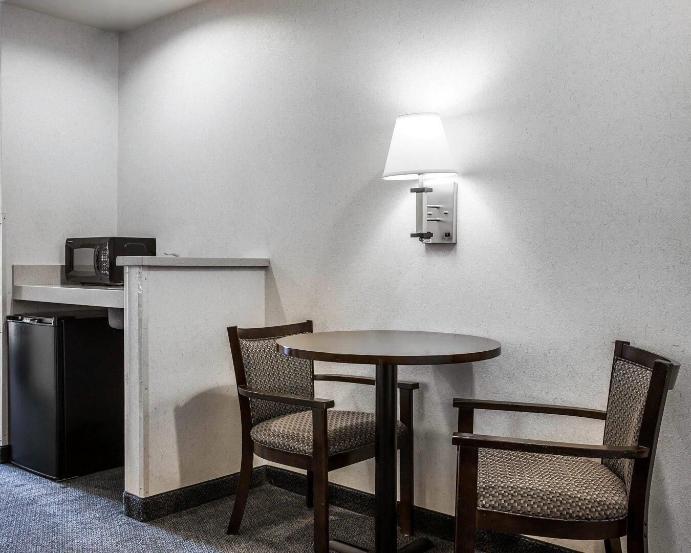 A small kitchenette with a microwave, refrigerator, and a wall-mounted light fixture. There are two chairs and a round table in the room.