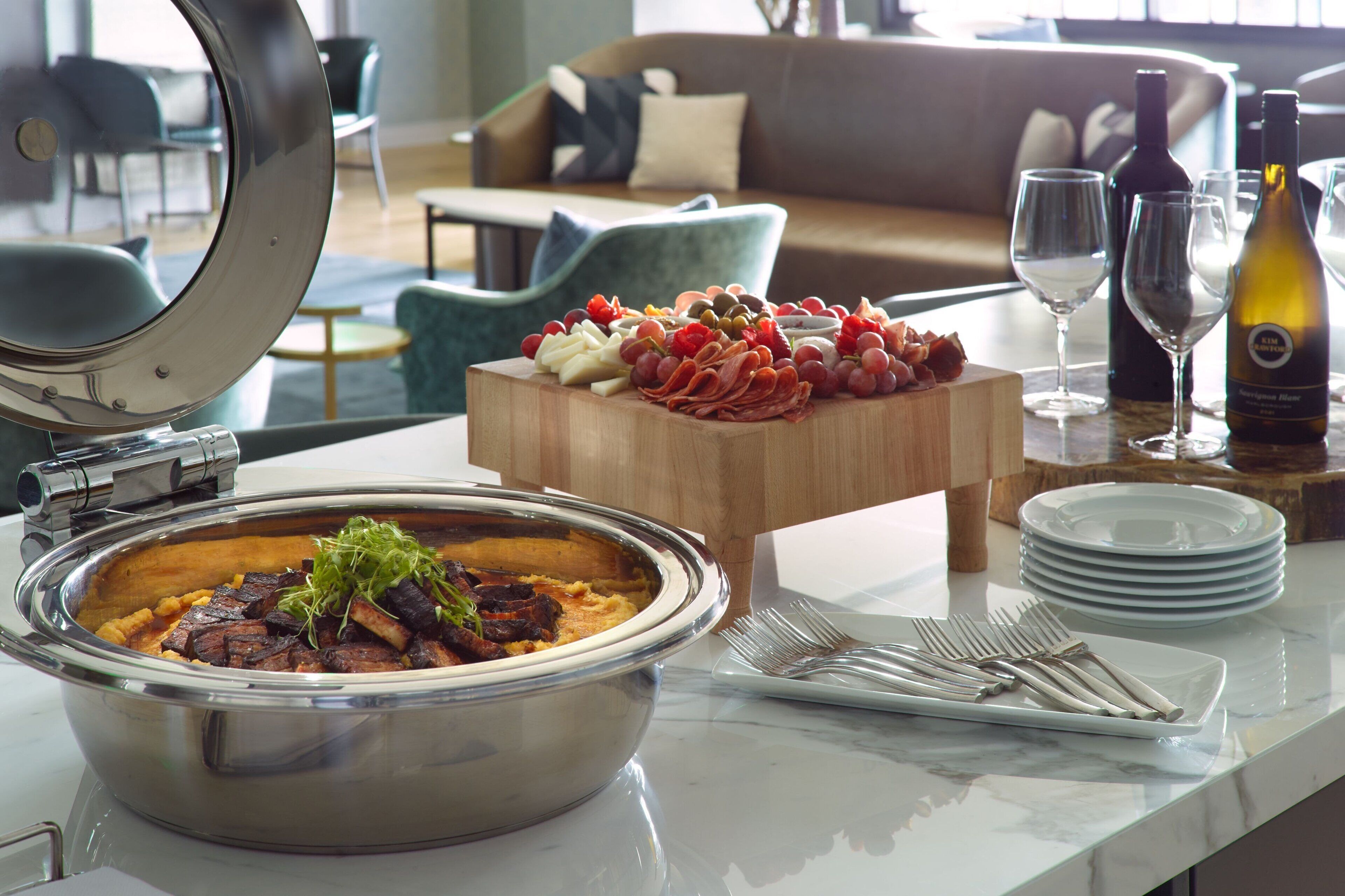 A buffet setup with a variety of foods, including a large bowl of soup, a wooden board with meats and cheeses, and a bottle of wine.