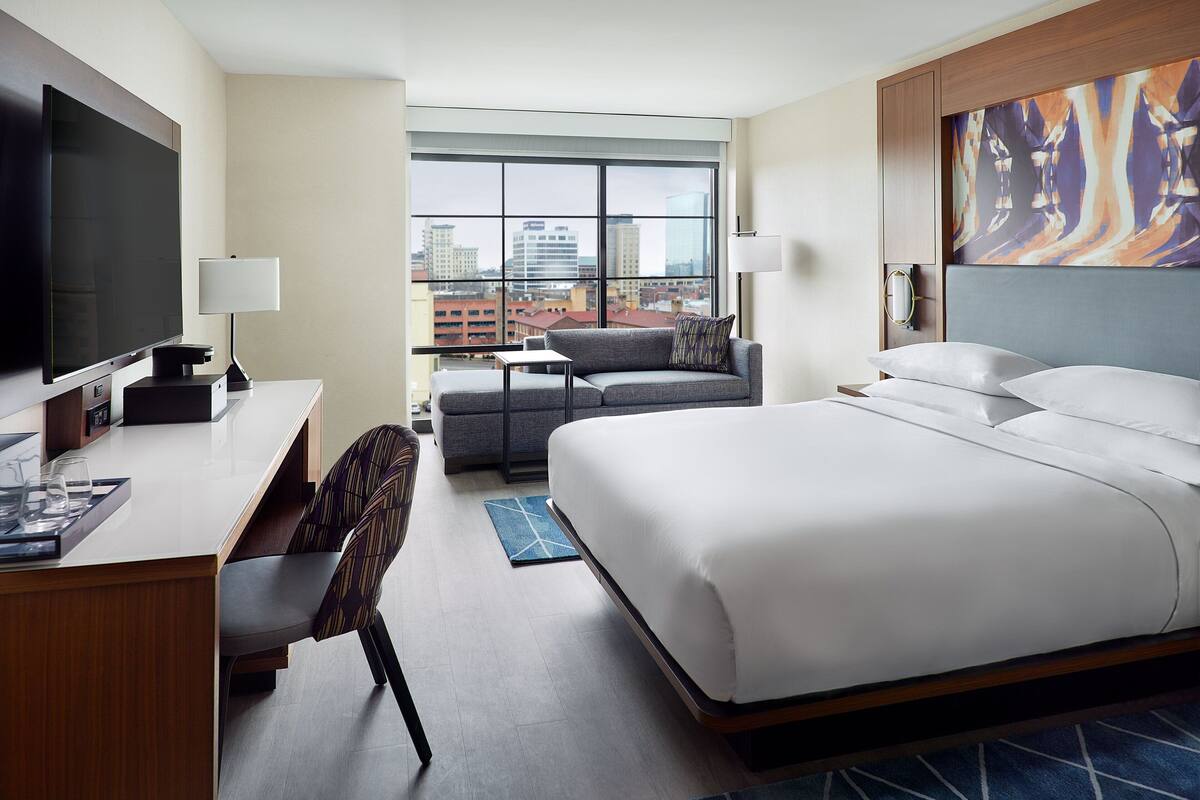 A hotel room with a large bed, a desk with a TV, a sofa, and a city view through large windows.