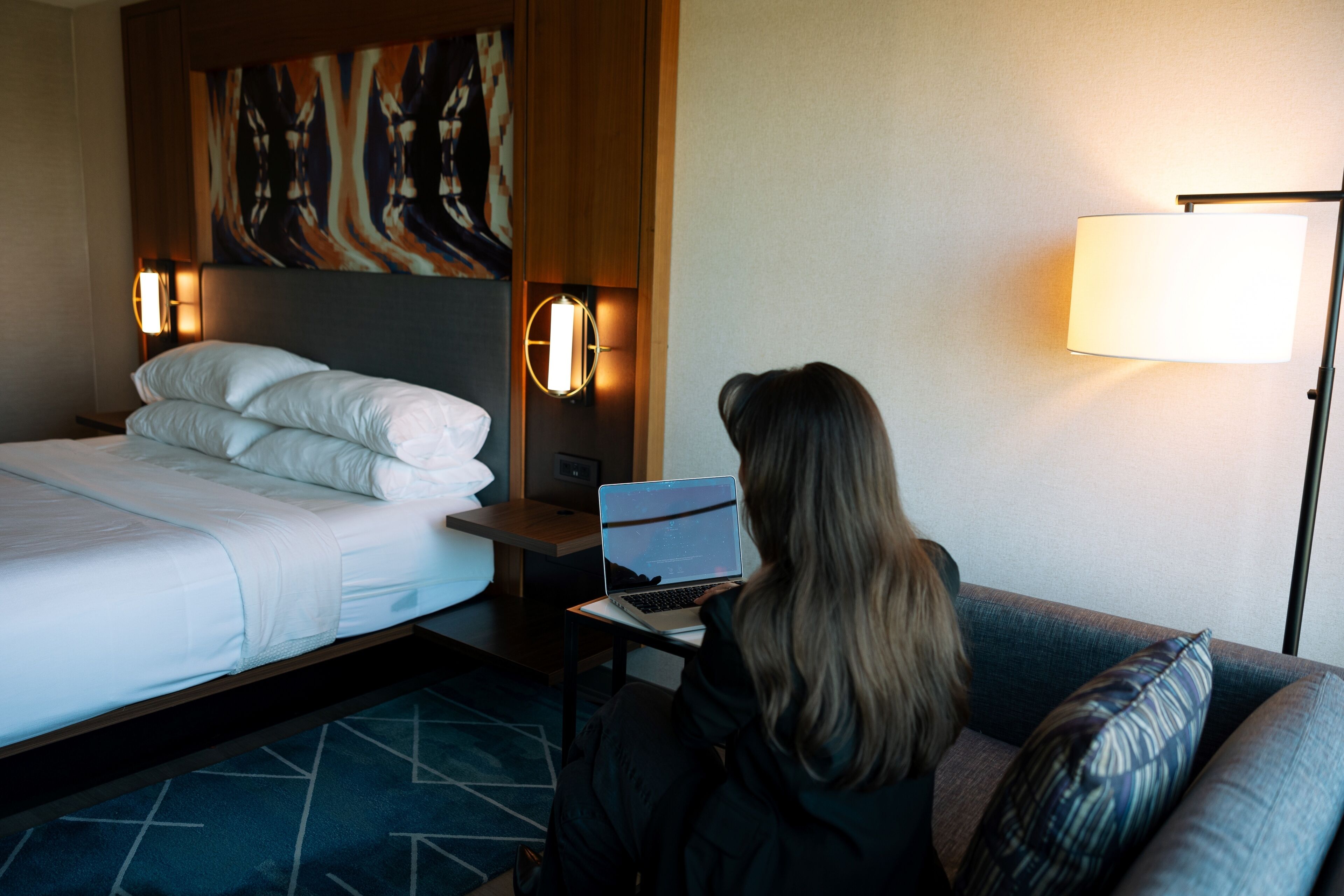 Een hotelkamer met een bed, een bureau met een laptop, een lamp en een schilderij aan de muur.