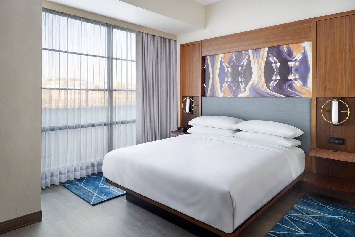 A modern hotel room with a large bed, a view of the city, and a wooden headboard.