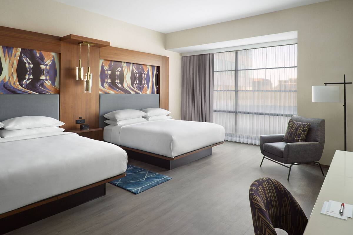 A hotel room with two beds, a desk, a chair, and large windows with curtains.