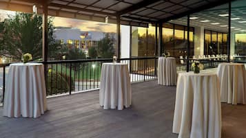 A covered outdoor area with round tables set up for dining, large windows offering views of the surrounding area, and a wooden floor.