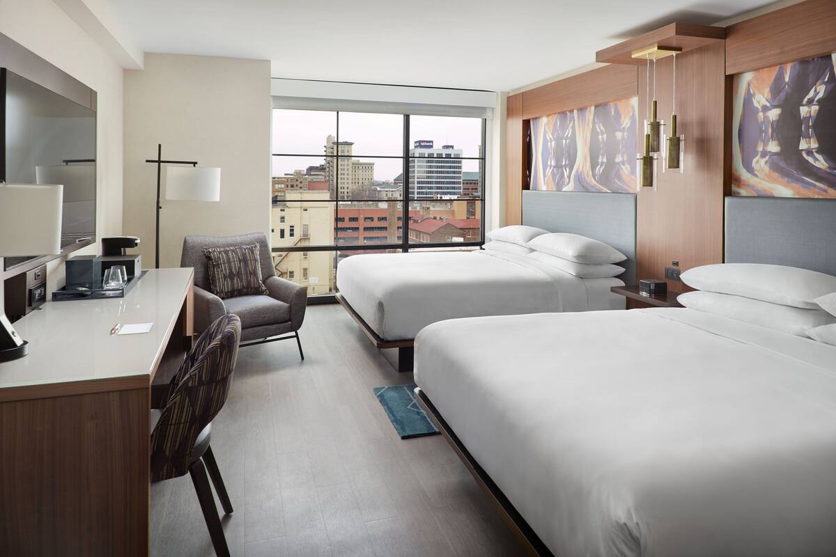 A hotel room with two beds, a desk with a computer, a chair, a lamp, and a large window offering a city view.
