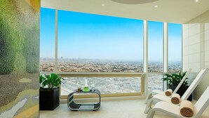 Ein Hotelzimmer in einem Hochhaus mit großen Fenstern und Blick auf die Stadt, einer kleinen Sitzecke mit Massagesessel und einer Dekoration an der Wand.