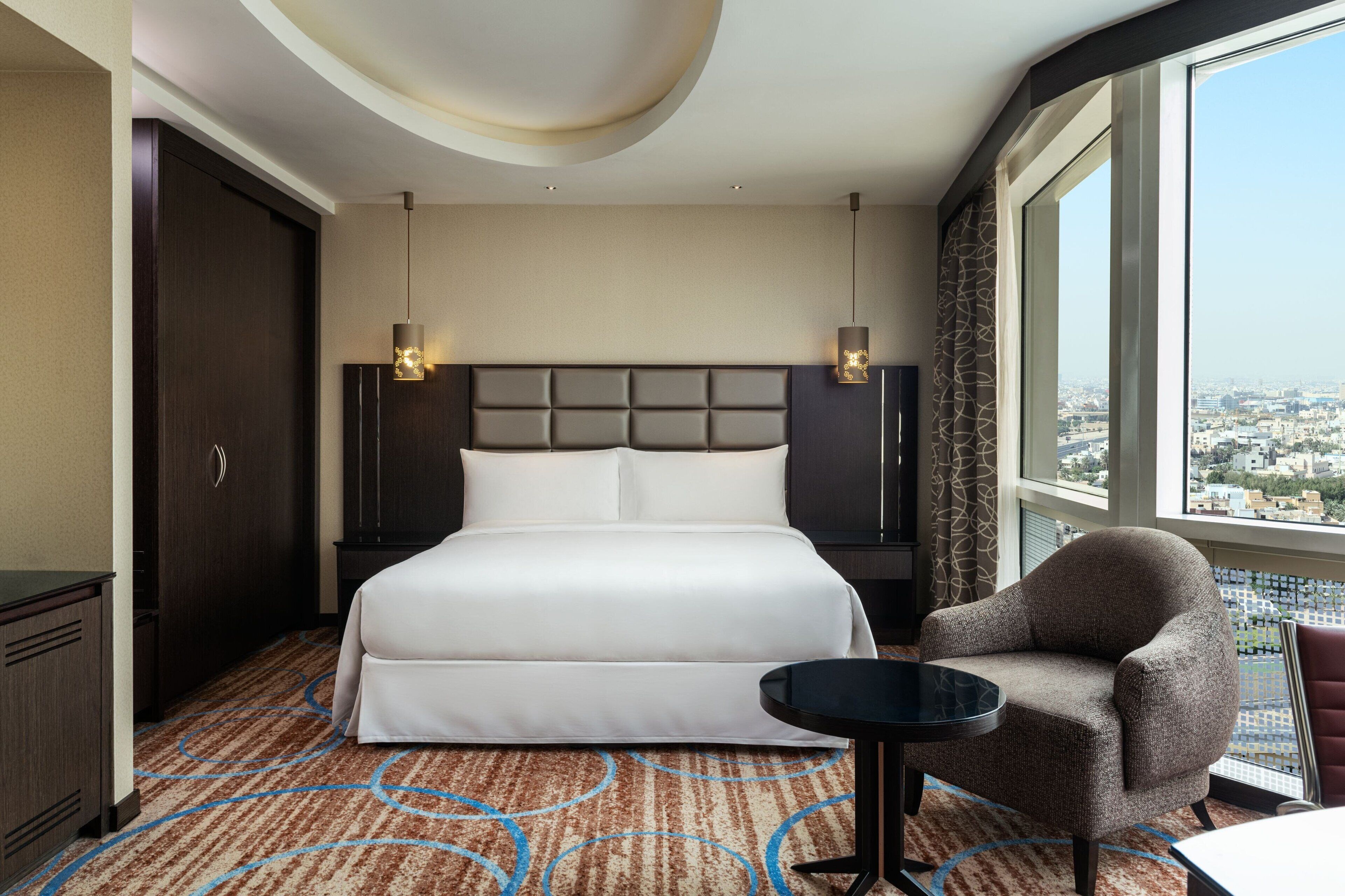 Une chambre d’hôtel avec un grand lit, une chaise, une petite table et une vue sur la ville par une grande fenêtre.