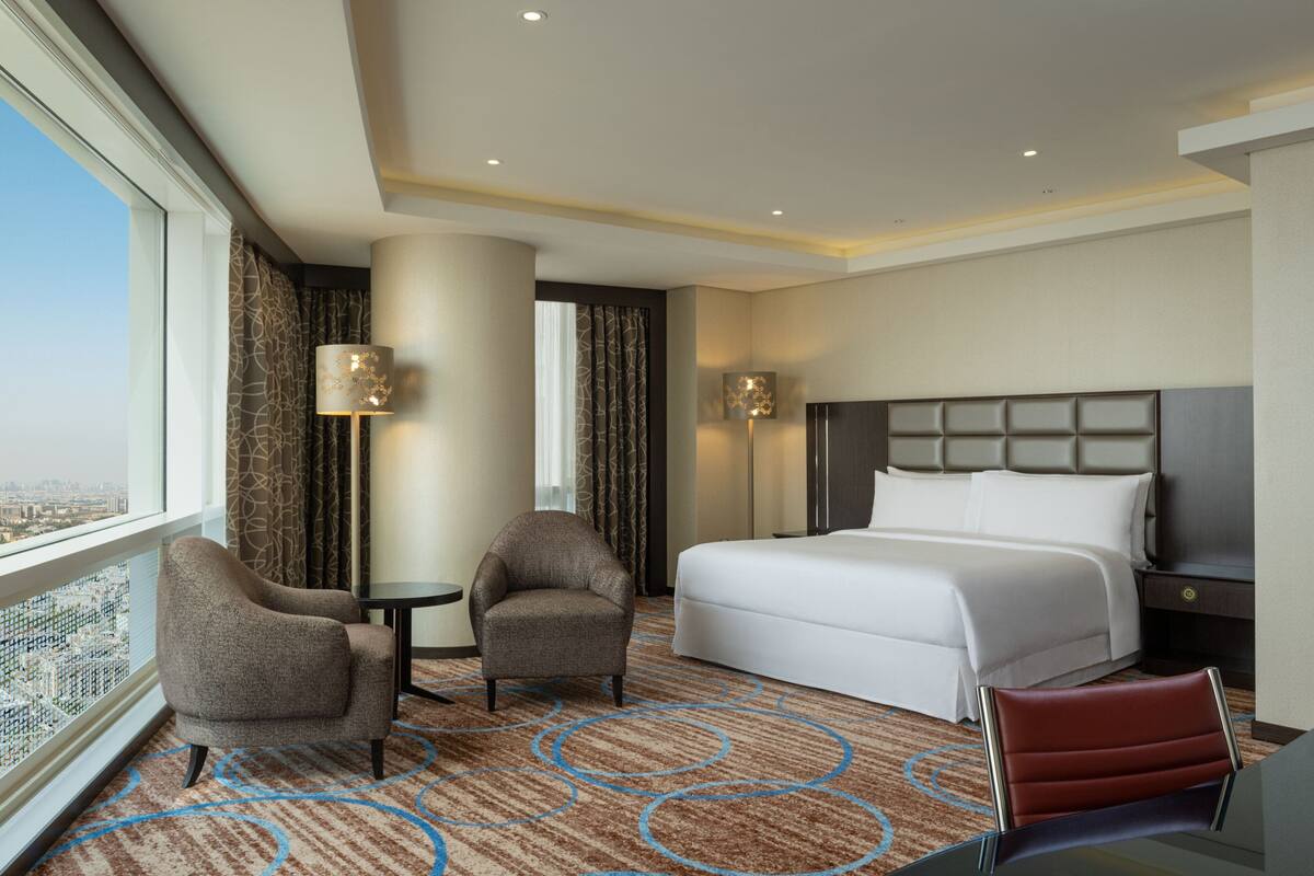 A hotel room with a large bed, two armchairs, a small table, and a view of the city.