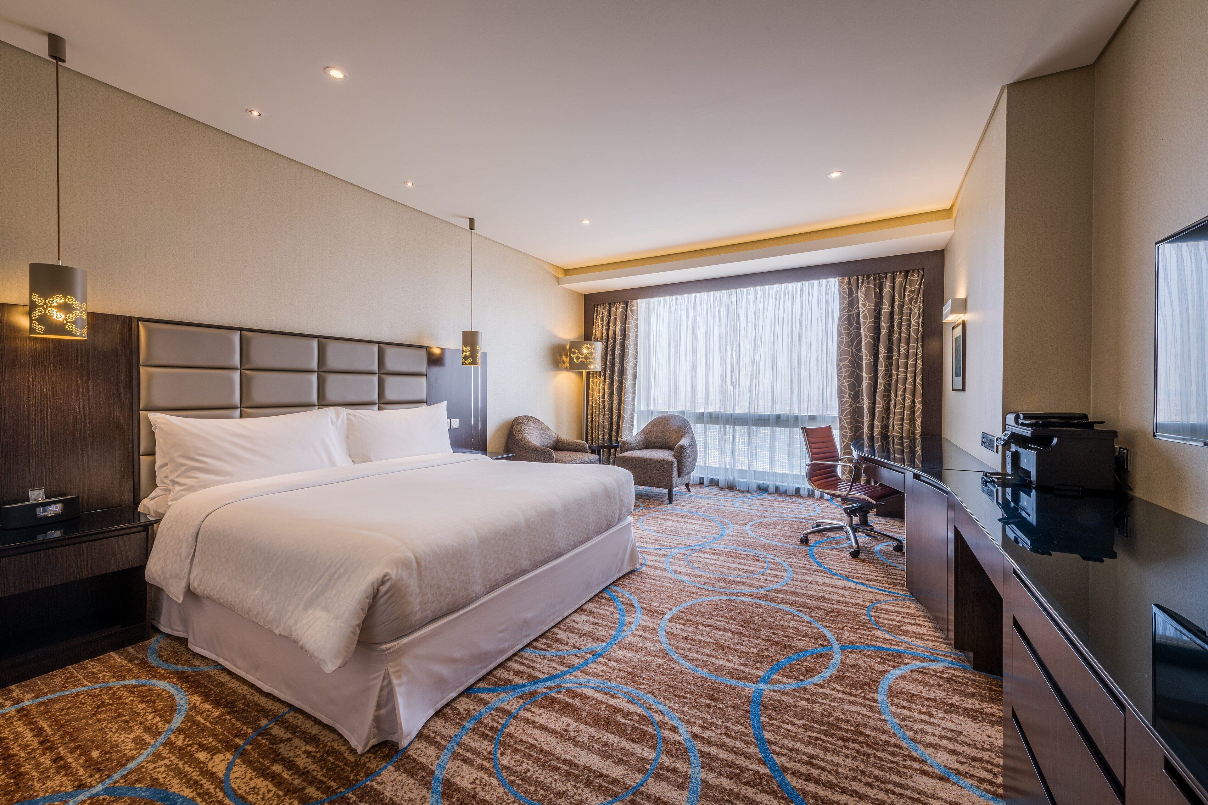 A hotel room with a large bed, a desk, a chair, a TV, and a view of the outdoors.