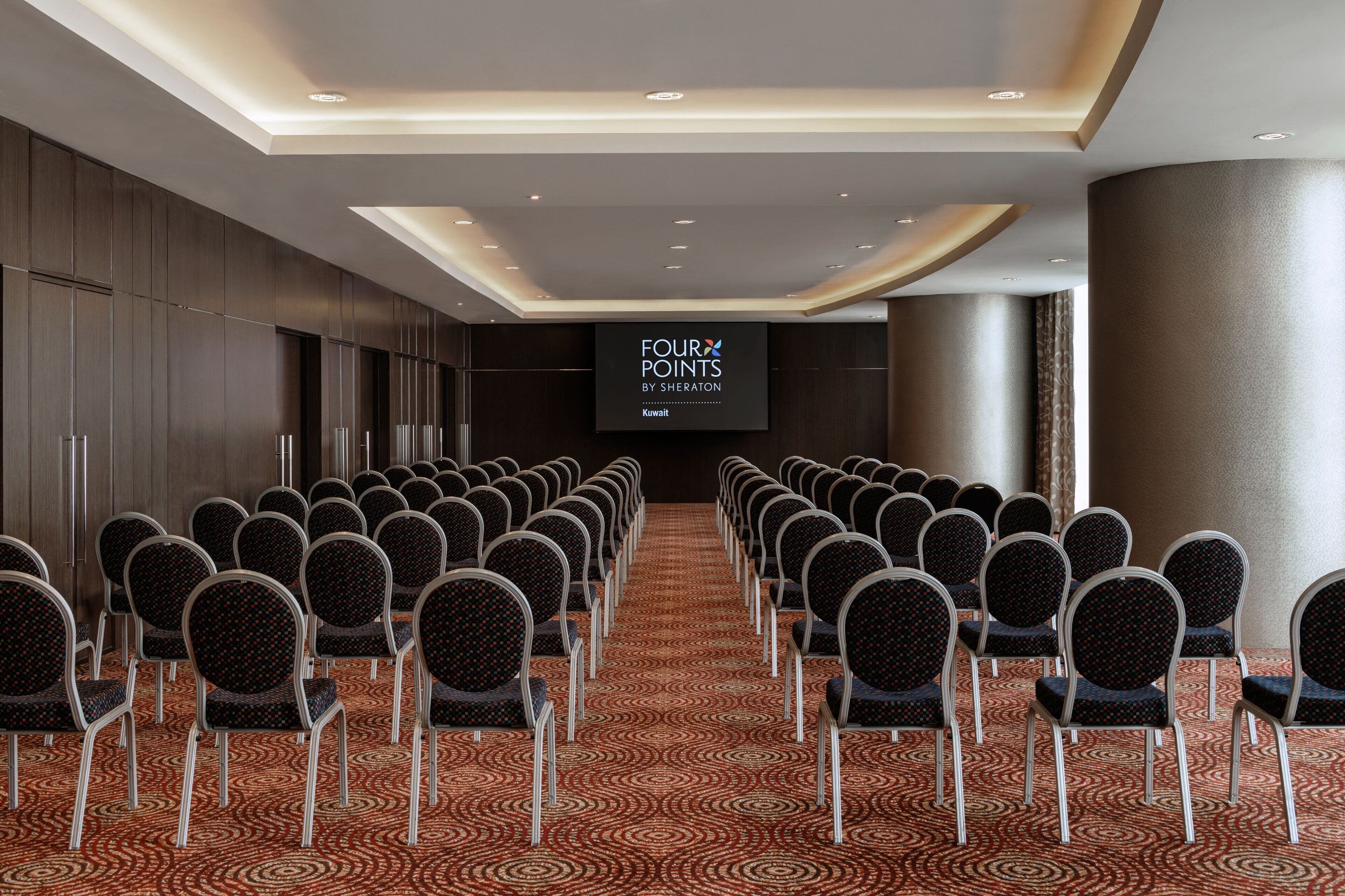 A conference room with rows of chairs, a large screen displaying "FOUR POINTS BY SHERATON Kuwait", and a patterned carpet.