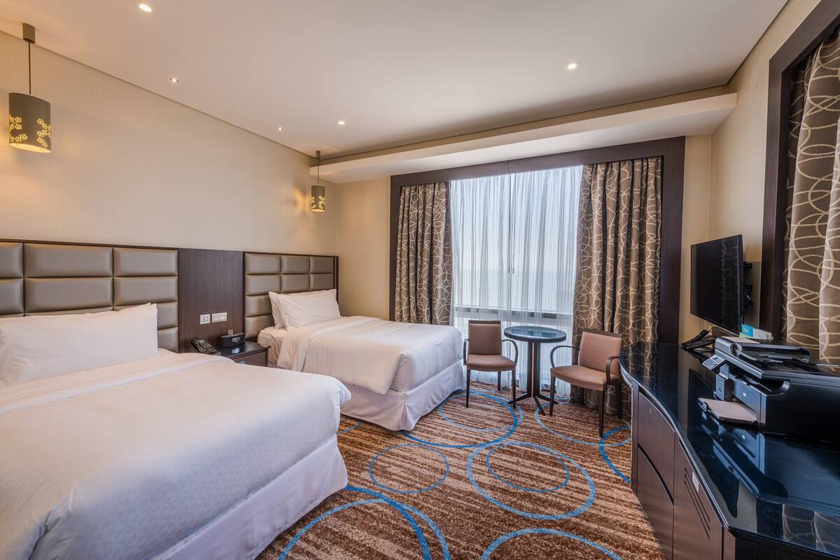 A hotel room with two beds, a desk with a printer, a chair, a TV, and a large window with curtains.