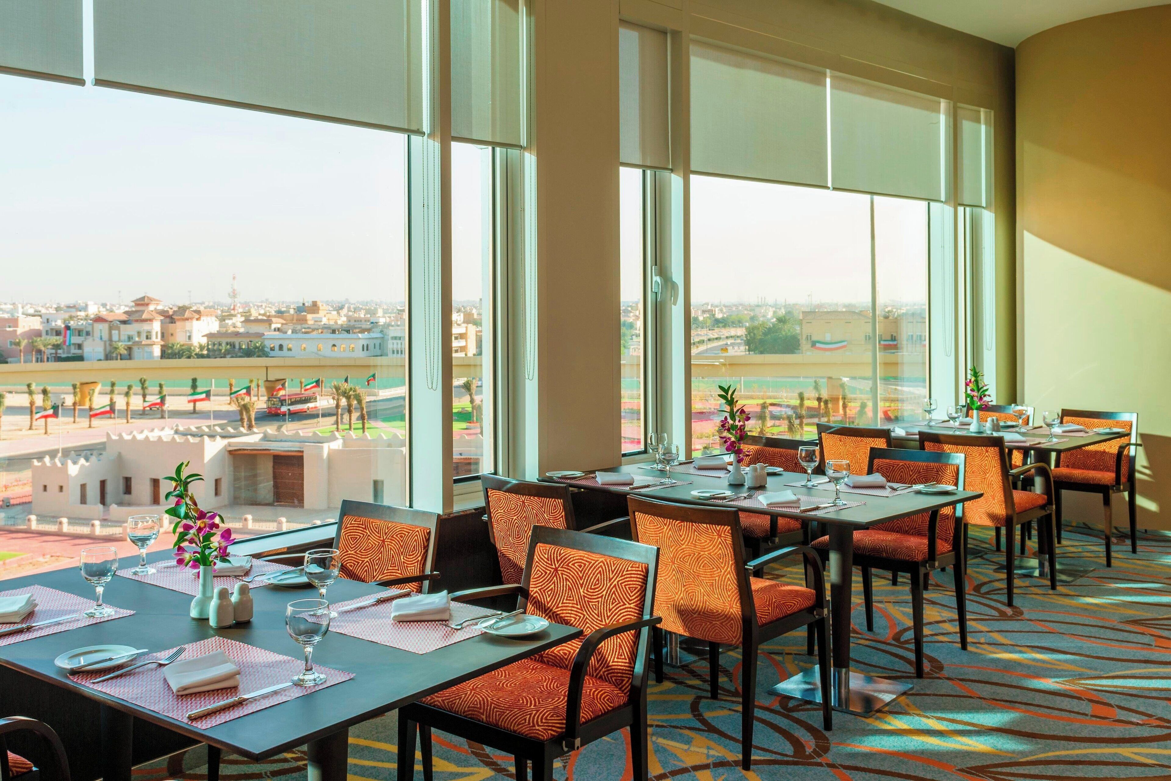 A dining area with tables set for four, large windows offering a view of the cityscape, and a patterned carpet.