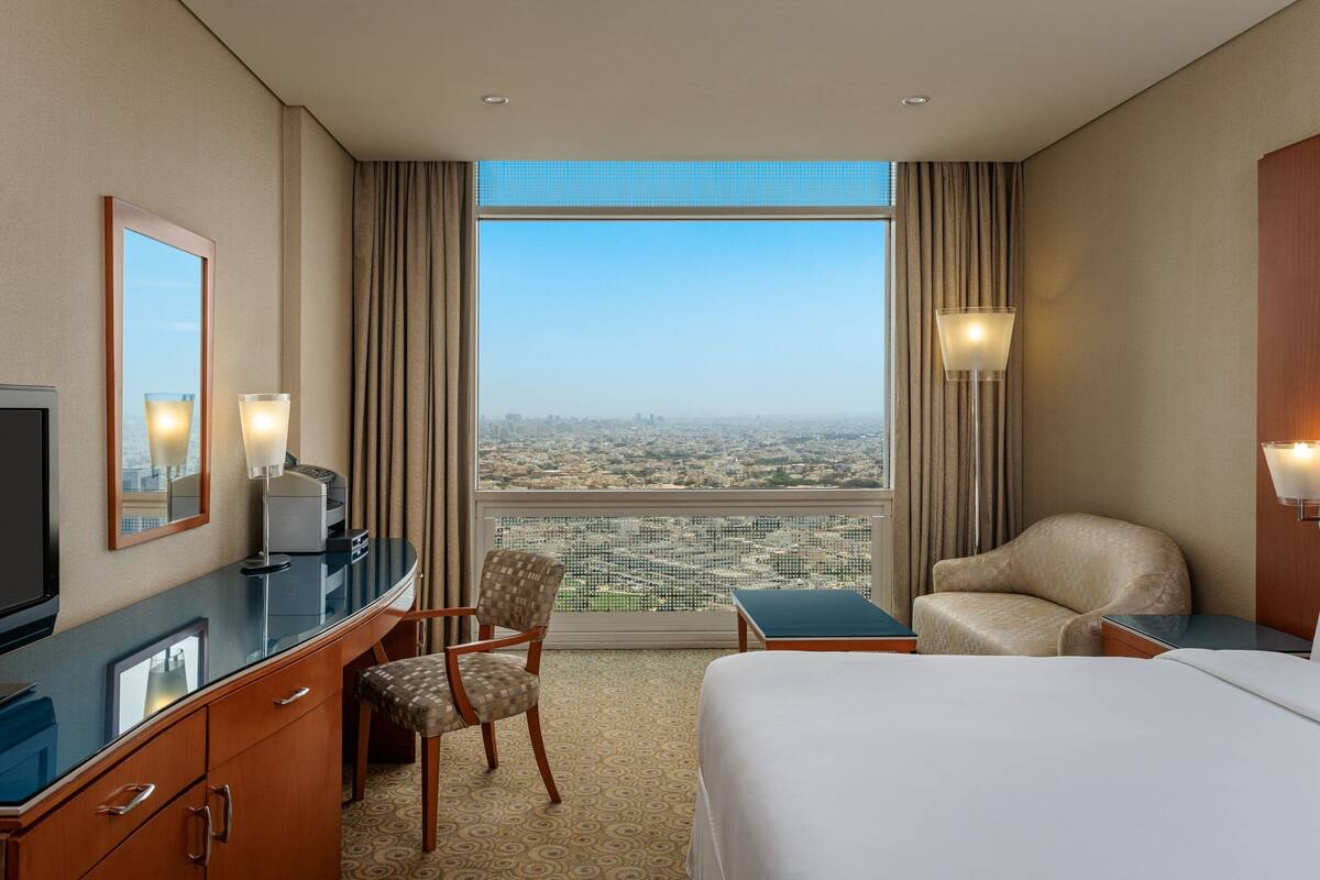 A hotel room with a large bed, a desk with a computer, a chair, a TV, and a view of a cityscape through a large window.