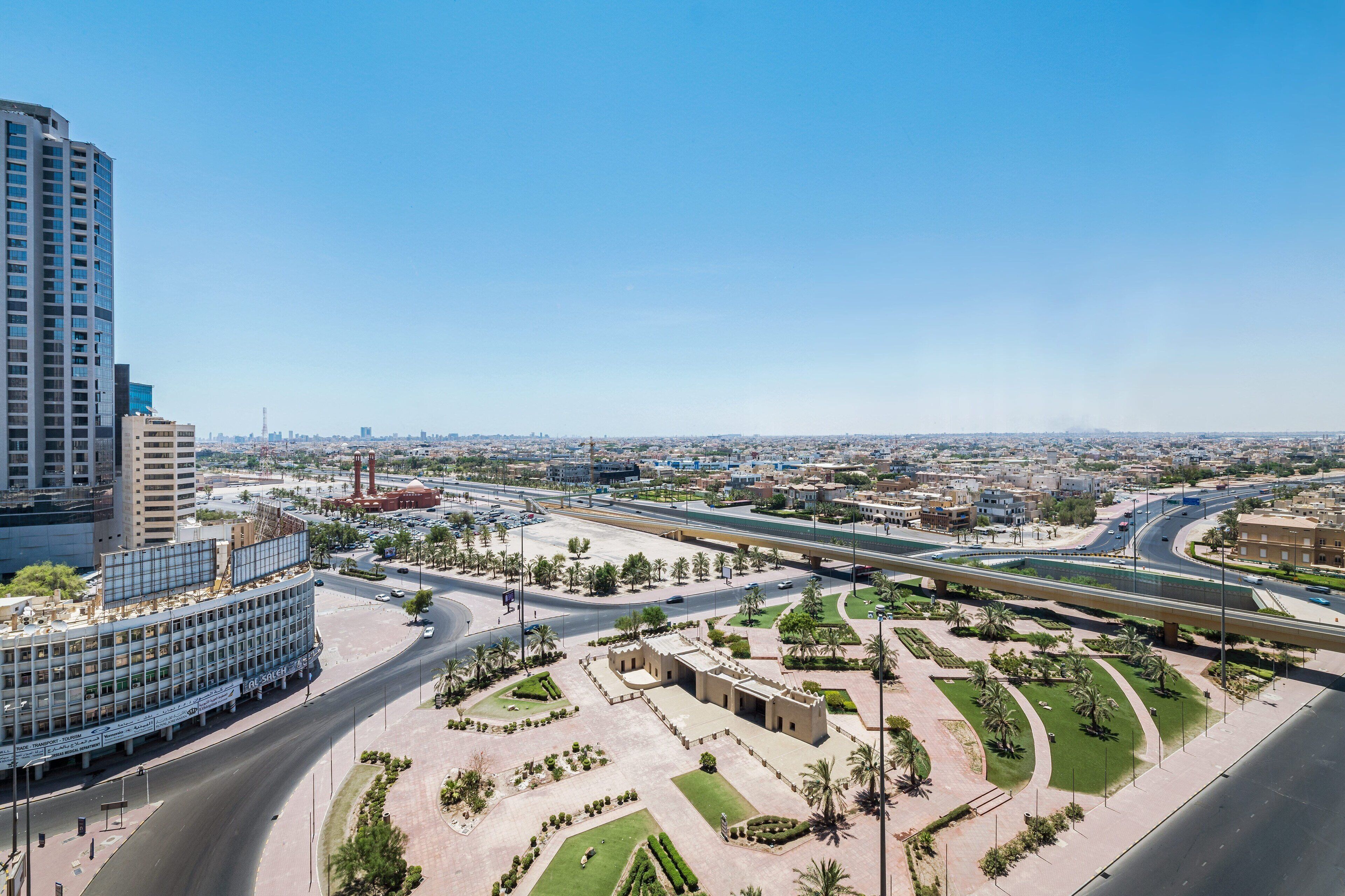 A modern urban landscape with a large building, a curved road, and a landscaped area with palm trees.