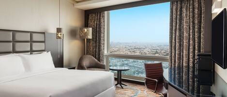 Habitación de hotel con una cama grande, un escritorio con silla, televisión y vista a la ciudad por la ventana.