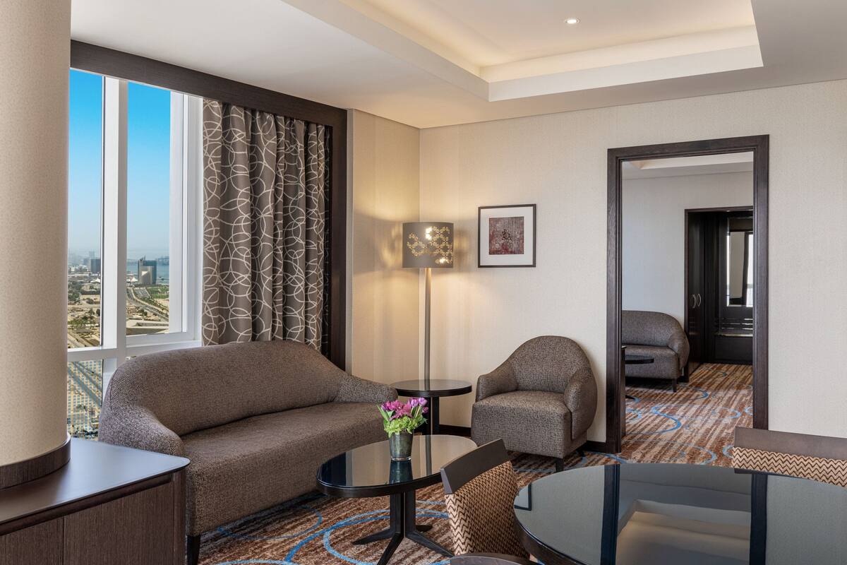 A hotel room with a sofa, armchair, small table, and a lamp. There is a large window with curtains, a framed picture on the wall, and a carpeted floor.