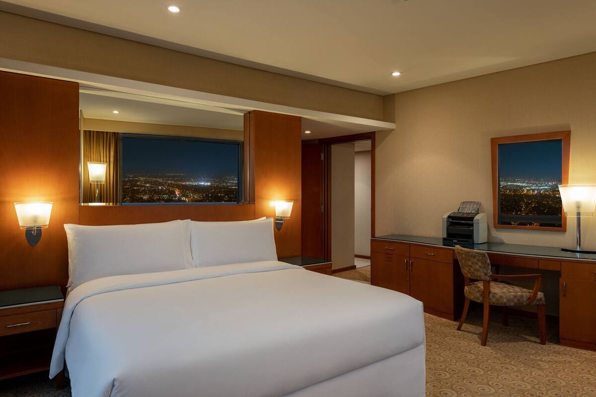 A hotel room with a large bed, a desk with a chair, a desk lamp, a window with a city view, and a mirror.
