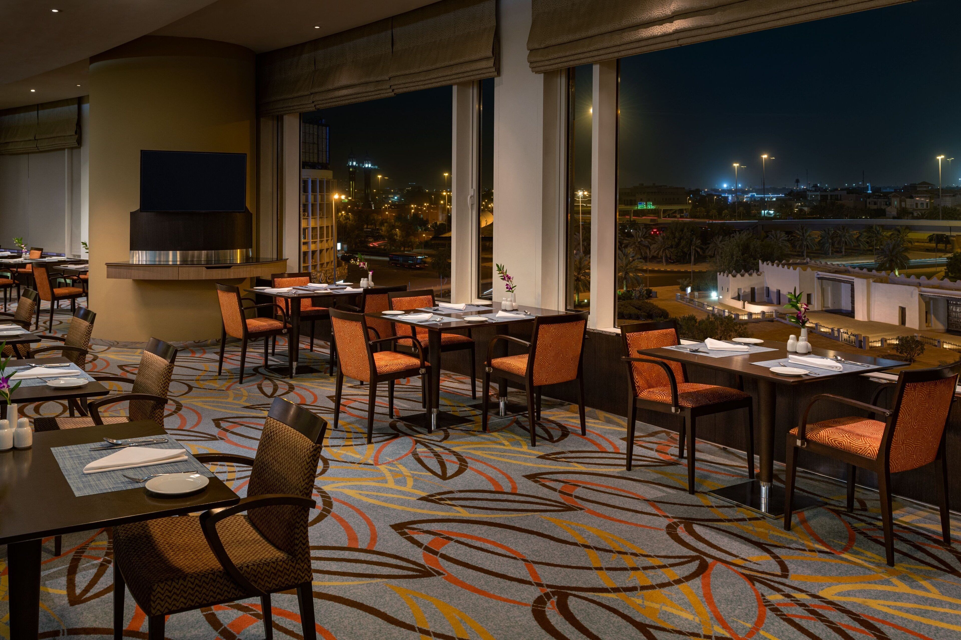A restaurant with tables and chairs, large windows, and a view of the city at night.