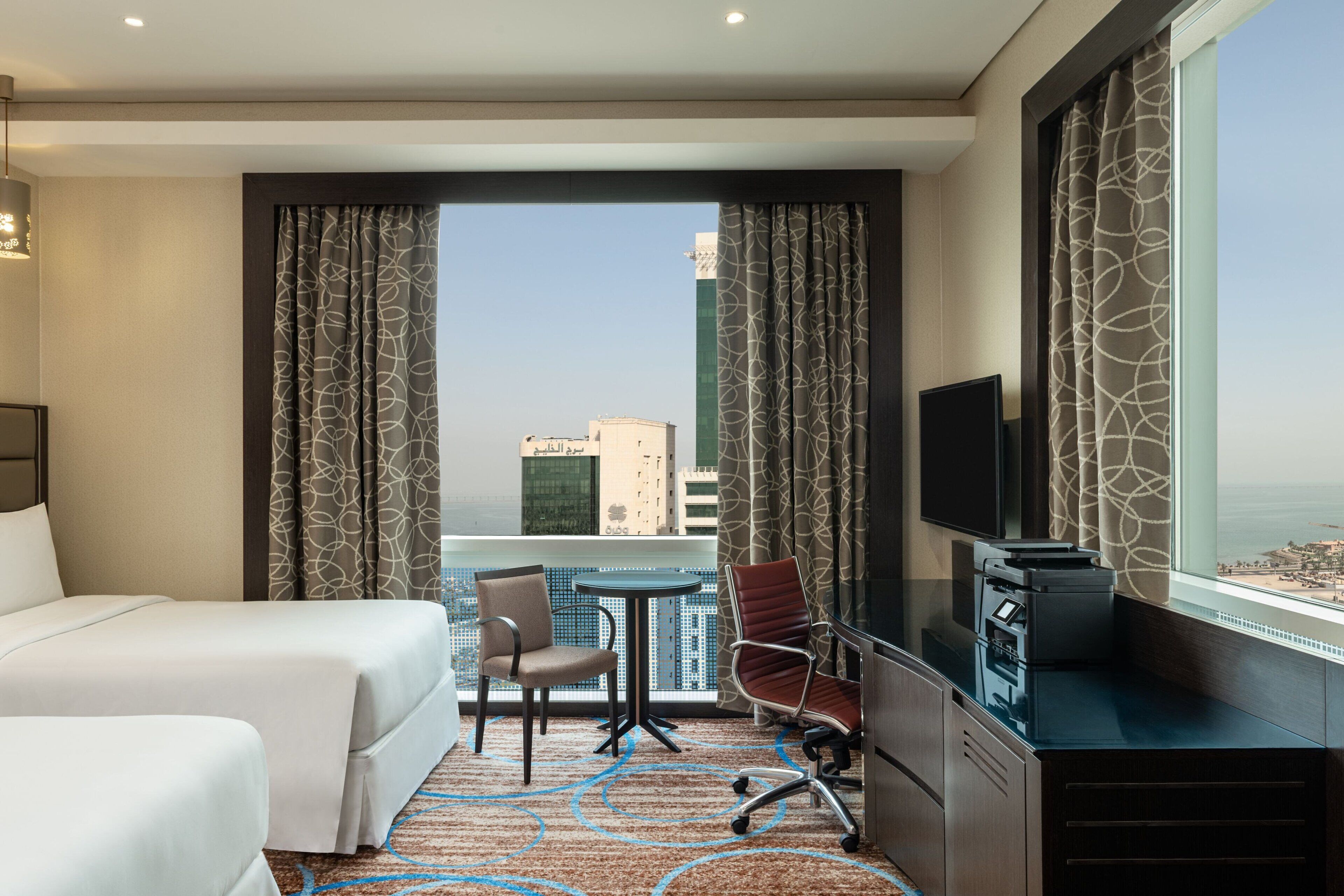 Une chambre d’hôtel avec un lit, un bureau, une chaise et un téléviseur. La fenêtre offre une vue sur les bâtiments et le paysage urbain.