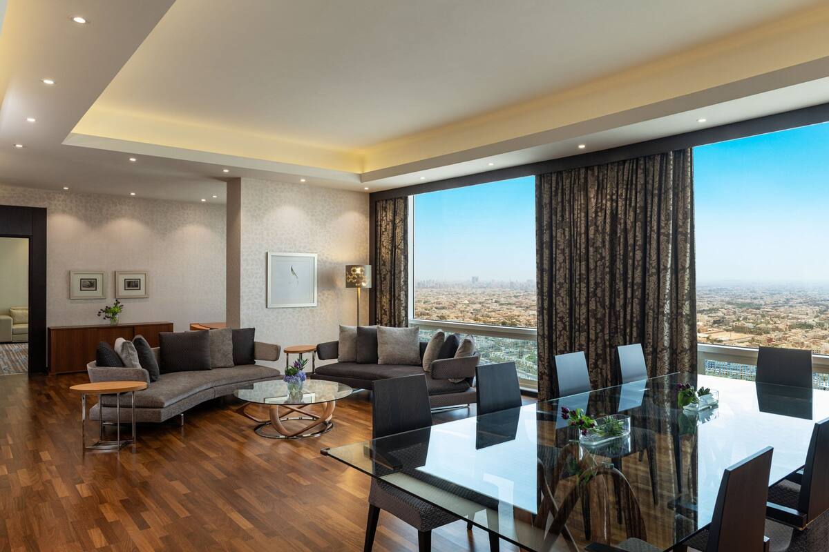 A modern living room with a glass dining table, sofas, and a large window with a view of the city.