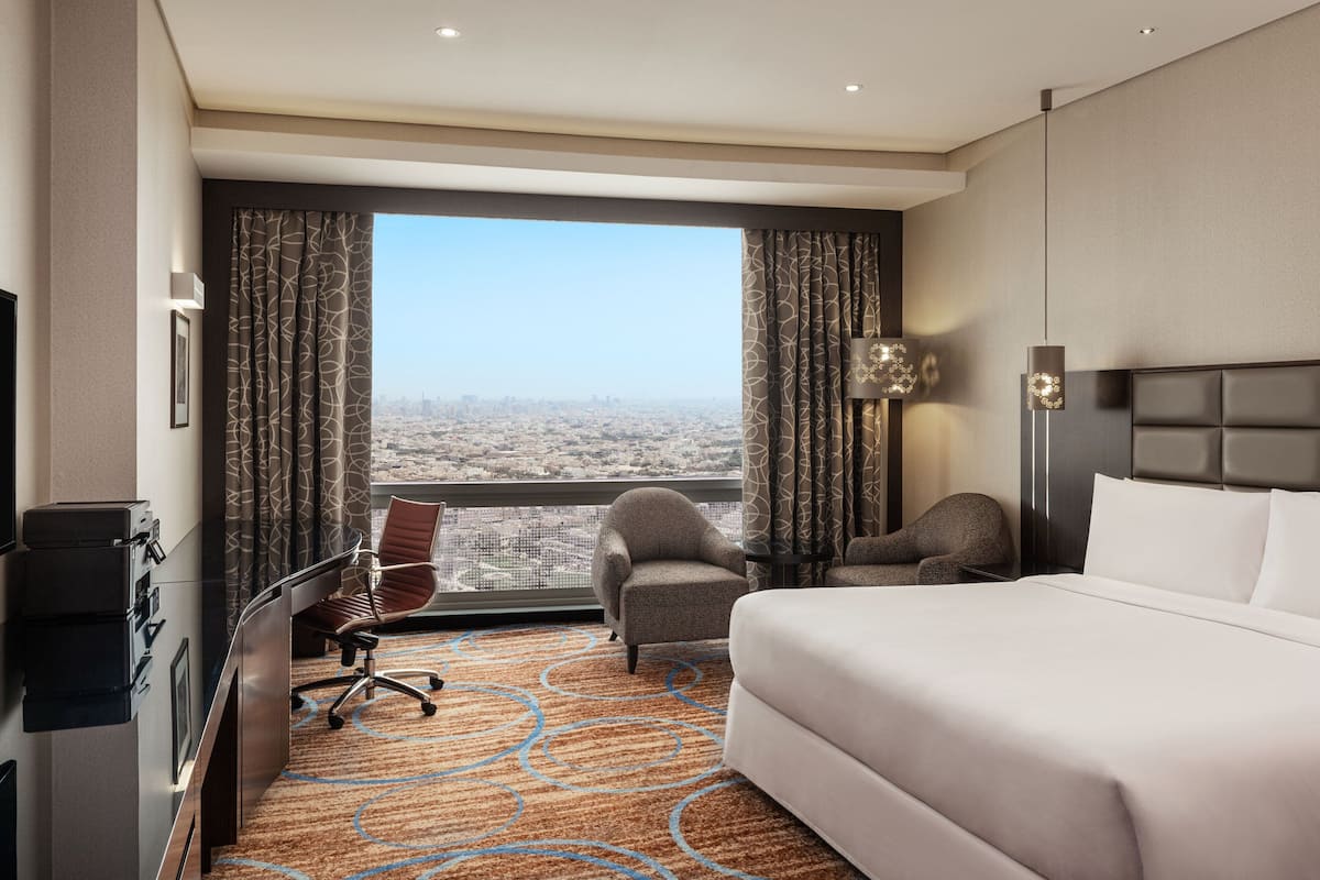 A hotel room with a large bed, a desk with a chair, a view of the city through large windows, and a modern lamp.