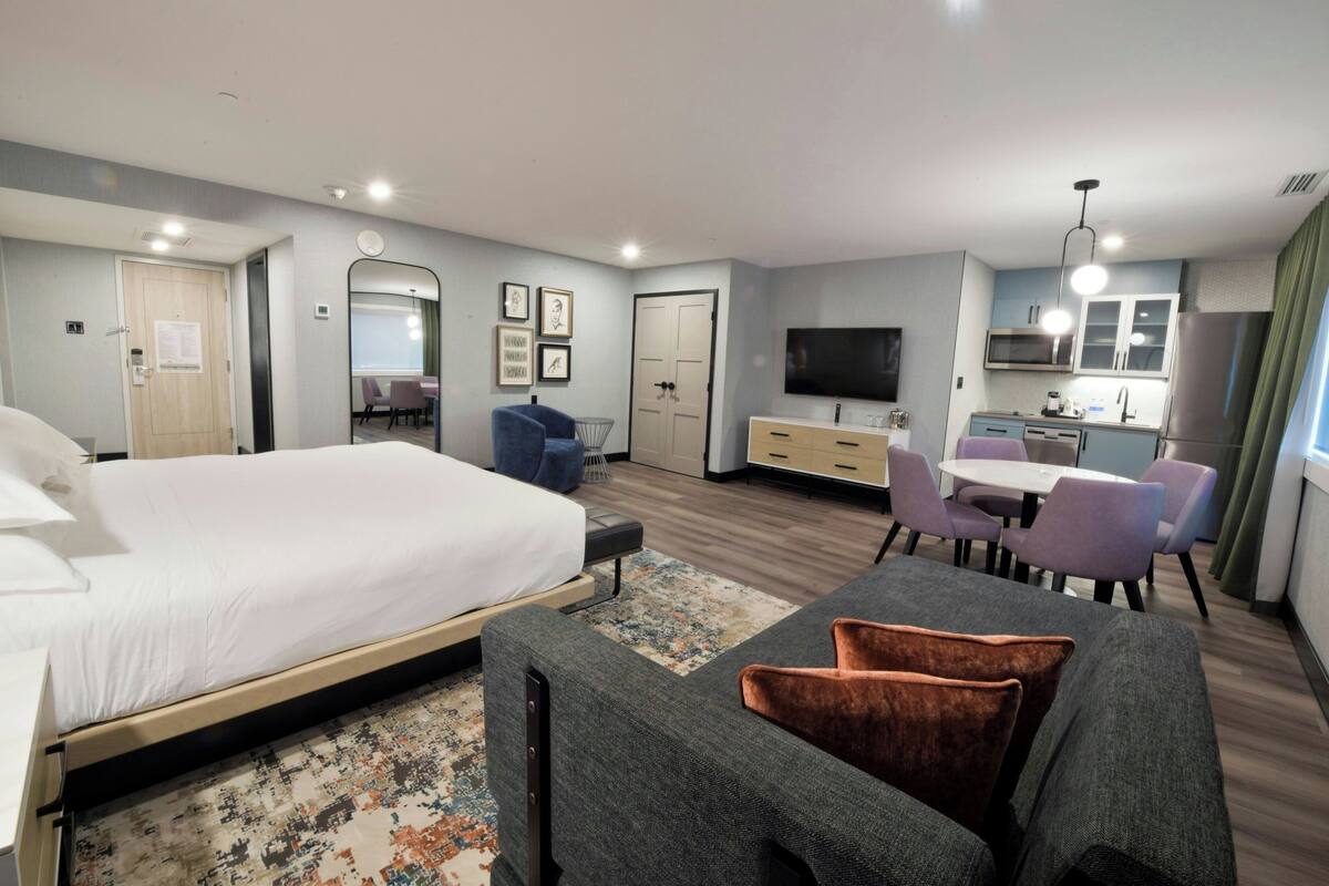 A hotel room with a bed, a desk, a chair, a TV, and a kitchenette.