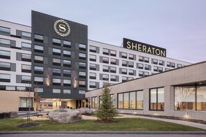 Un hôtel Sheraton à l’architecture moderne, doté d’une façade élégante, de grandes fenêtres et d’un espace extérieur bien entretenu, agrémenté de rochers et d’un petit arbre.