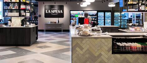 A modern café interior with a herringbone tile floor, a central counter with a coffee machine, and shelves stocked with various food items.